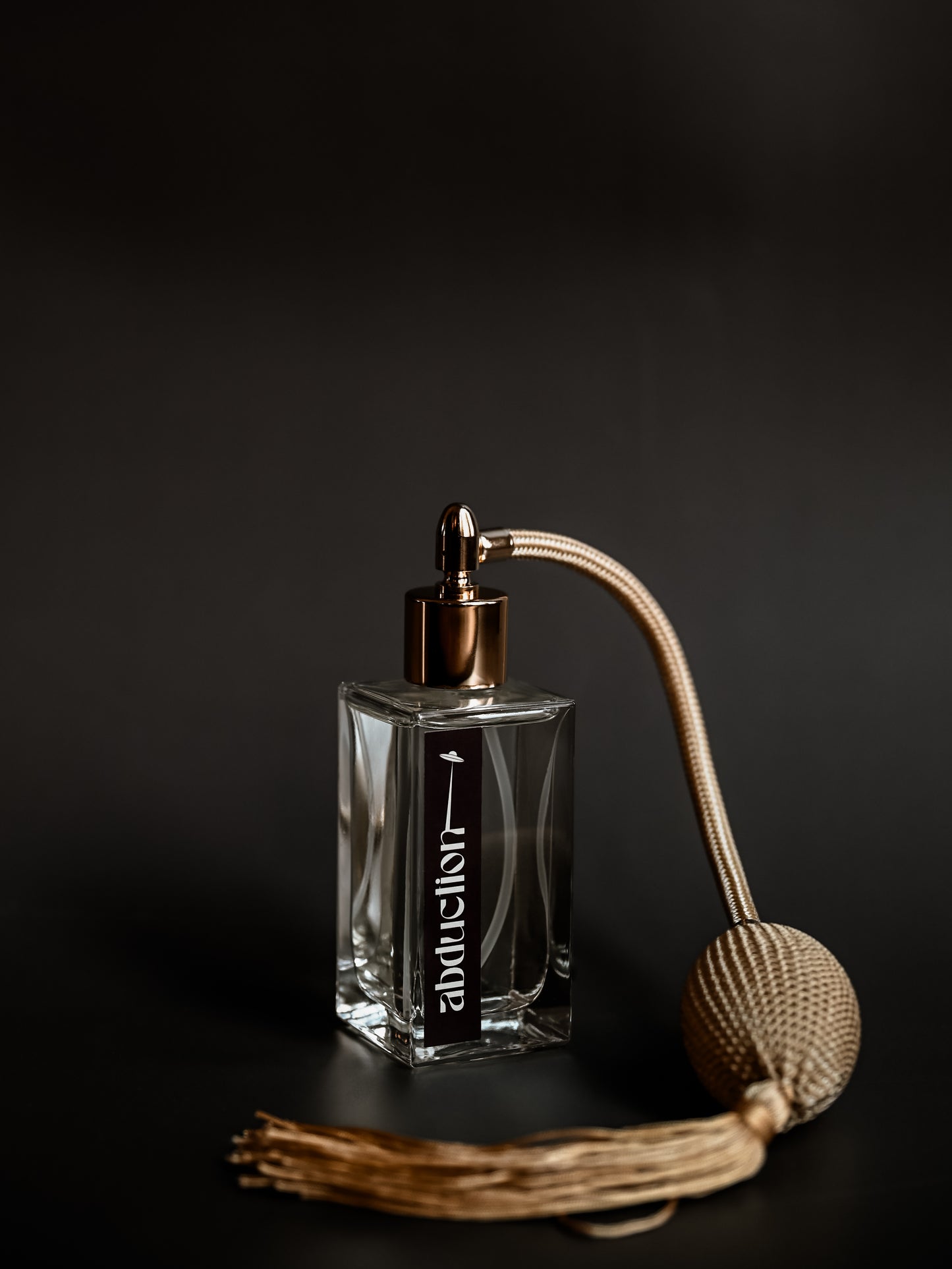 A clear glass perfume bottle with a vintage-inspired design, featuring a metallic cap and a bulb sprayer with a tassel. The label reads 'Abduction'.