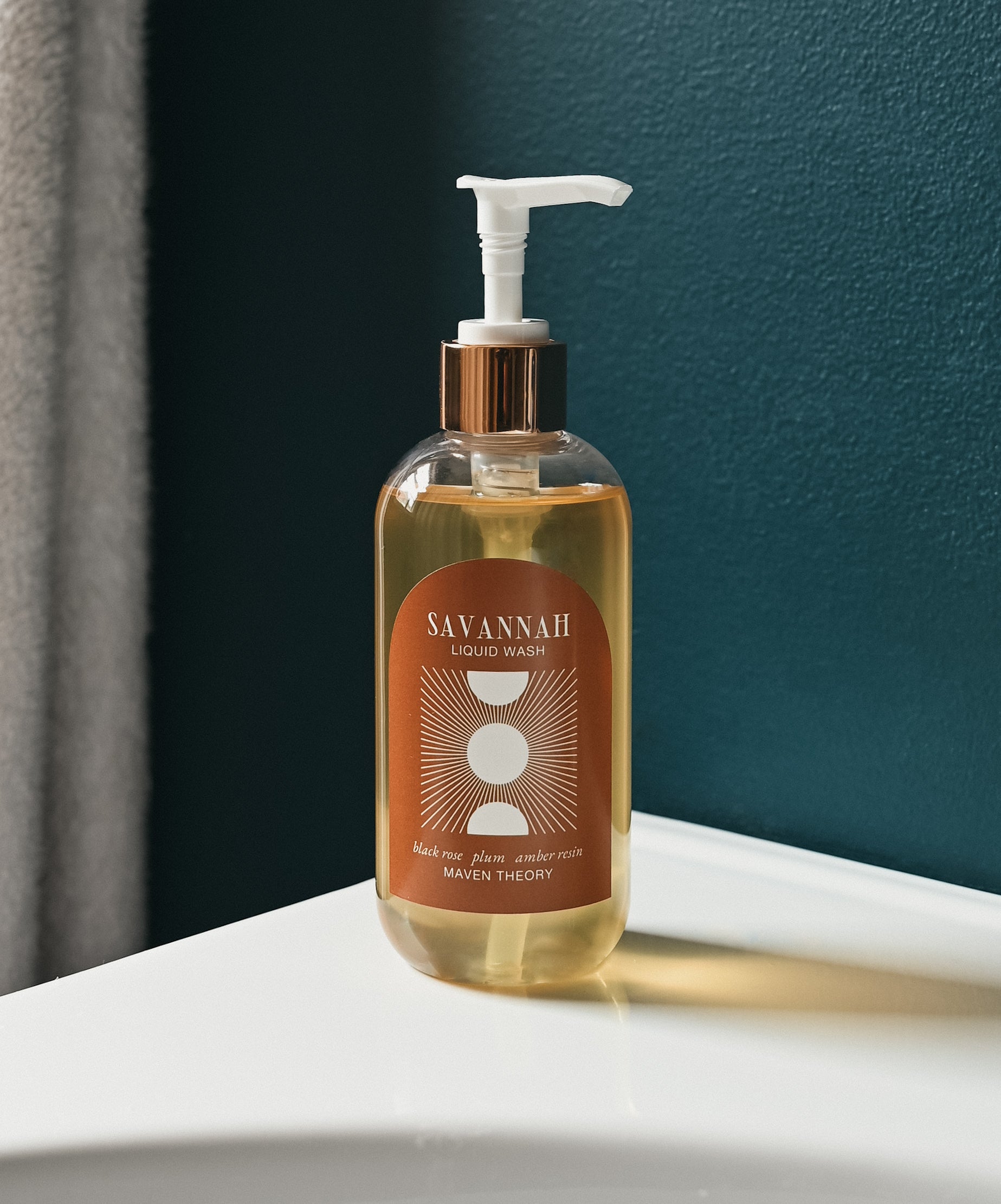 A clear pump bottle of Savannah hand & body wash with a bronze label, placed on a white surface with a dark blue wall in the background.